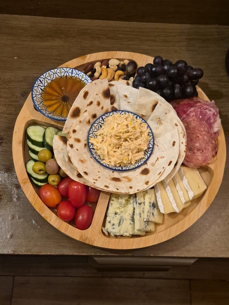 Tábua redonda com queijos, salame, uvas, castanhas, tomates, pepino, azeitonas e pães folha
