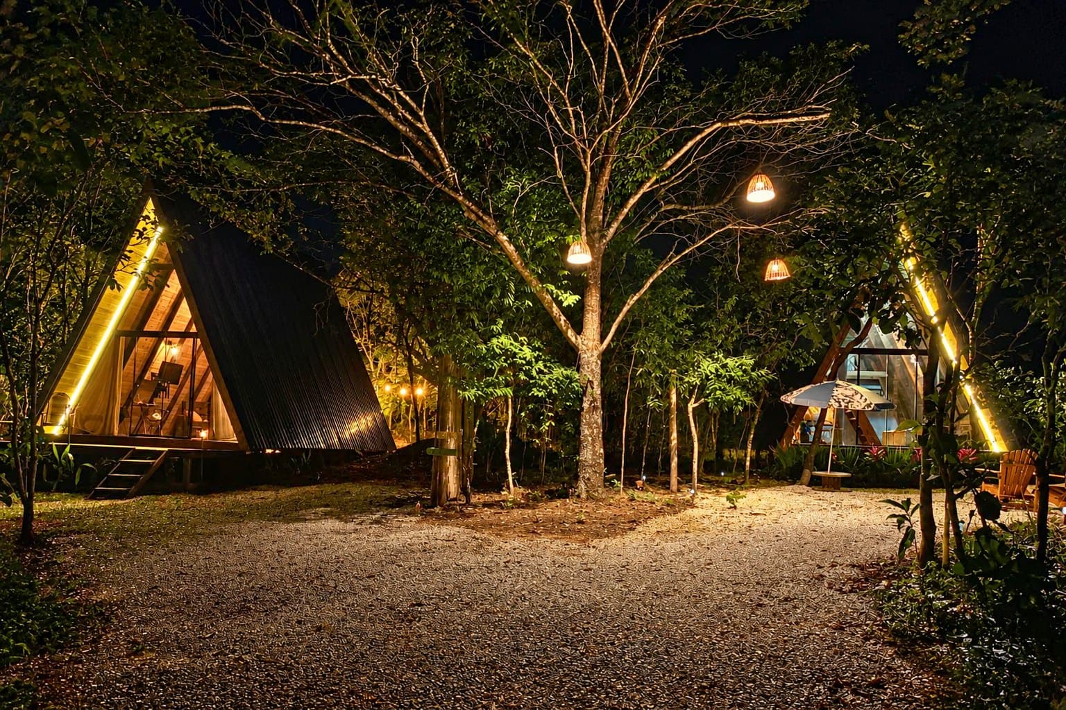 Os dois chalés triangulares iluminados à noite, entre as árvores, com luminárias penduradas e caminho de cascalho
